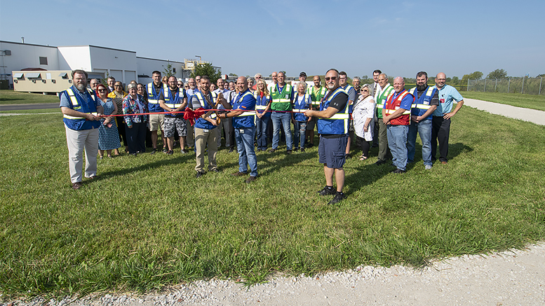 Community leaders, local officials, and Magna Powertrain leadership are pictured during ribbon-cutting ceremonies held today. Pictured holding the scissors for the ribbon-cutting are Stephen Brand, Magna Powertrain General Manager and Joe Barr, Assistant General Manager. The walking track for employees can be seen winding around the property. Photo by: Mike Rhodes