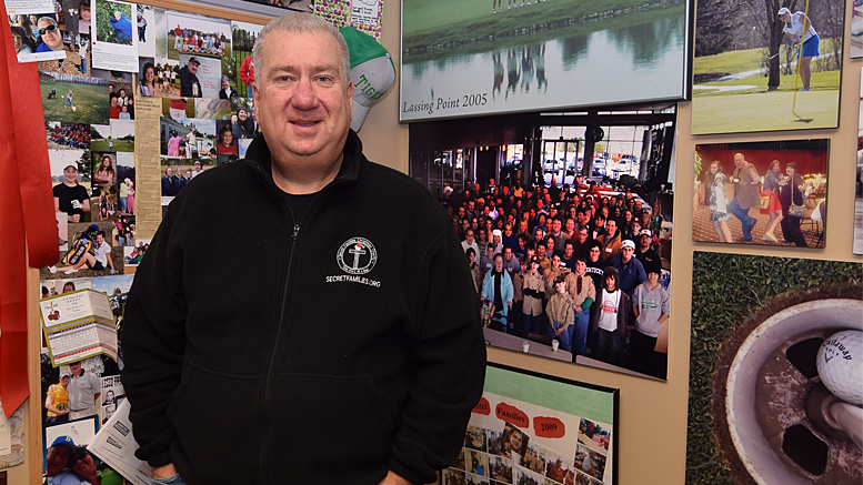 Al Holdren is pictured in his office standing beside hundreds of photos of thousands of volunteers who participate in "Secret Families." Photo by: Mike Rhodes